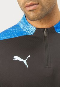 Schwarzes Sportoberteil mit einem Viertel-Reißverschluss, strukturiertem blauen Kragen und einem reflektierenden weißen Puma-Logo auf der Brust. Glattes Material mit kontrastierenden Farben.