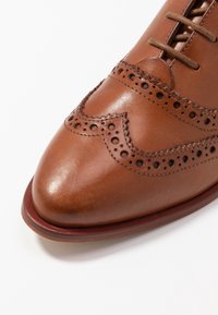 Chaussure en cuir marron avec des détails en brogue, bout pointu et lacets marron. Présente des accents perforés et une texture lisse.