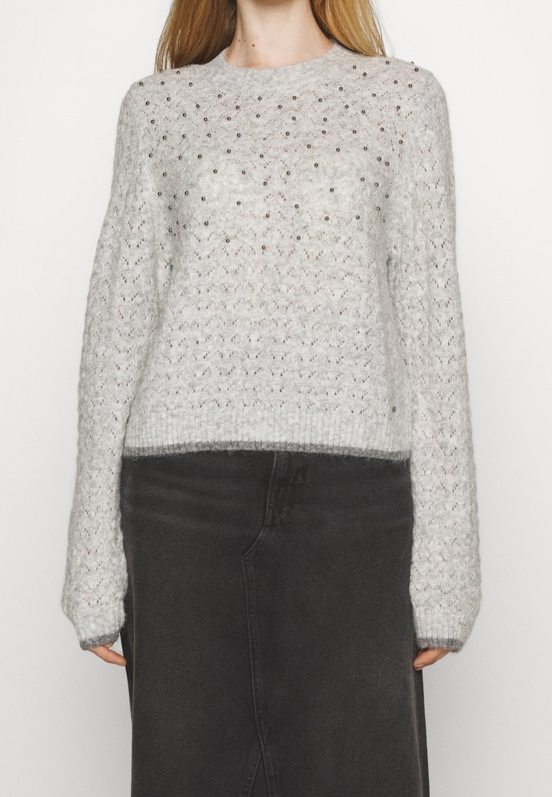 Pull tricoté gris clair avec de petites décorations en perles sur la poitrine et manches longues, associé à une jupe foncée taille haute.