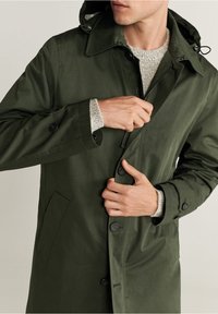 Oliivinvihreä takki hupulla, jossa on vetoketju, nappikiinnitys ja sivutaskut. Kangas näyttää sileältä ja kevyen tuntuiselta.