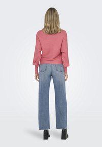 Frau mit schulterlangem blonden Haar, die einen rosa gerippten Pullover, blaue weite Jeans und schwarze Stiefeletten mit Absatz trägt, mit dem Rücken zur Kamera stehend.