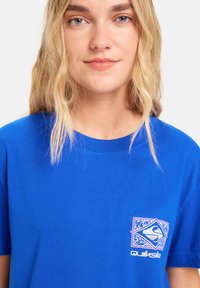 Blaues Baumwoll-T-Shirt mit Rundhalsausschnitt, kurzen Ärmeln und einem weißen und rosa Quiksilver-Logo auf der linken Brust. Weicher Stoff und lockere Passform.