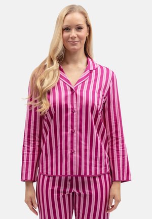 Frau mit langen blonden Haaren, die einen Pyjama mit leuchtend pinken und hellrosa vertikalen Streifen trägt, steht vor einem einfarbigen Hintergrund.