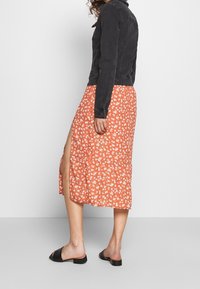 Kvinna som bär en svart denimjacka över en orange blommig midikjol med en sidospalt och svarta låga slip-on-sandaler, står på sidan.