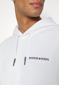 Bílá mikina s kapucí s přední klokankovou kapsou, šňůrkami s černými konci a logem "SCOTCH & SODA" ve černé výšivce.