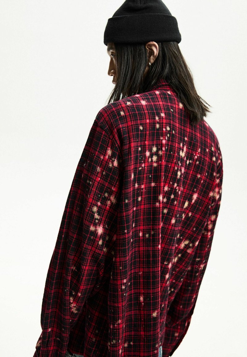 Chemise à carreaux rouge et noir avec une coupe décontractée, présentant un motif usé et des taches de décoloration légères. Le matériau semble être un tissu doux.