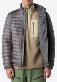 Pohodlná šedá bunda s podšívkou, která má zapínání na zip a boční kapsy, oblečená přes béžový fleecový svetr s krémovou kapsou na hrudi.