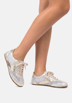 Piernas con zapatillas casuales blancas y plateadas metálicas de malla con cordones y detalles beige sobre un fondo claro y liso.