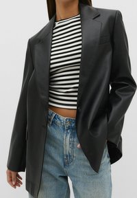 Vrouw in zwarte leren blazer, zwart-wit gestreepte crop top en lichtblauwe hoge taille jeans, hand in zak, tegen een effen achtergrond.