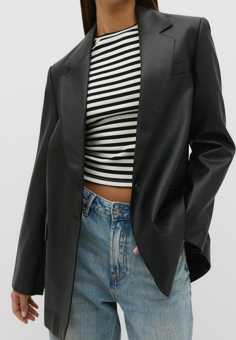 Vrouw in zwarte leren blazer, zwart-wit gestreepte crop top en lichtblauwe hoge taille jeans, hand in zak, tegen een effen achtergrond.