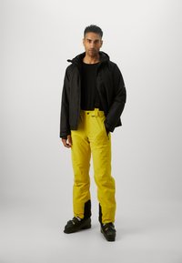 Jachetă neagră cu glugă, top negru casual, pantaloni galbeni strălucitori cu talie ajustabilă și cizme de schi negre, pe un fundal neutru.