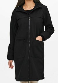 Mujer vestida con un abrigo negro con cremallera y capucha, con las manos en los bolsillos, de pie contra un fondo blanco liso.