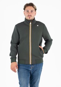 Donkergroen zip-up jack met hoge kraag, witte en oranje verticale rits en voorzakken. Gladde textuur en nauwsluitend ontwerp.