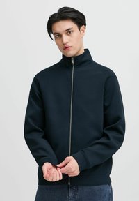 Marineblå zip-up jakke med høy krage og ribbede mansjetter. Laget av mykt stoff med en jevn tekstur. Har en rett silhuett og glidelåsdetaljer.
