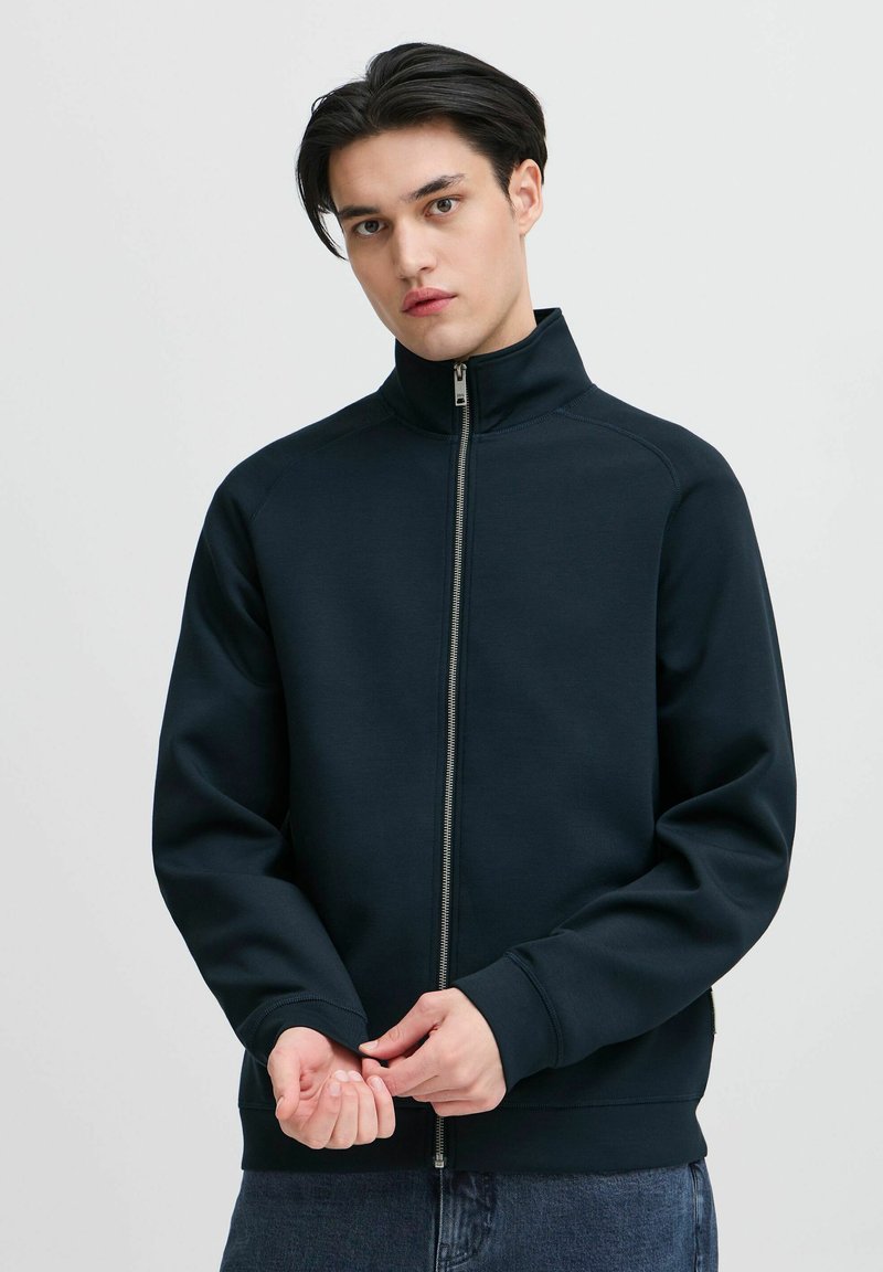 Marineblå zip-up jakke med høy krage og ribbede mansjetter. Laget av mykt stoff med en jevn tekstur. Har en rett silhuett og glidelåsdetaljer.