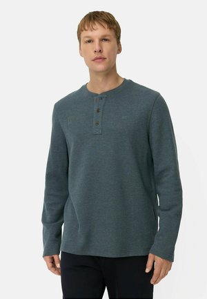 Junger Mann trägt ein dunkelgrünes Henley-Shirt mit langen Ärmeln und drei Knöpfen, steht vor einem schlichten hellen Hintergrund.