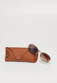 Rudos be rėmelio Ray-Ban saulės akinių su gradientiniais lęšiais, ilsinčiųsi ant atitinkamo rudo odinio Ray-Ban dėklo.