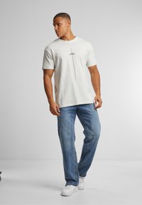 Light gray T-shirt with short sleeves and a central black vertical line design, paired with blue jeans and white sneakers.
