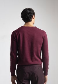 Suéter burdeos de manga larga, con cuello redondo y puños acanalados. Diseño de punto texturizado. Combinado con pantalones oscuros, mostrando un ajuste casual.