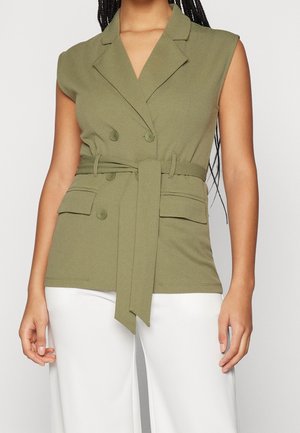 Blazer sans manches vert olive ceinturé avec revers et poches avant à rabat, porté avec un pantalon blanc.