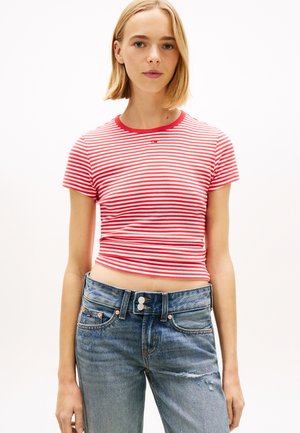 Jeune femme portant un t-shirt court à rayures rouges et blanches et un jean bleu taille haute déchiré, debout devant un fond blanc.