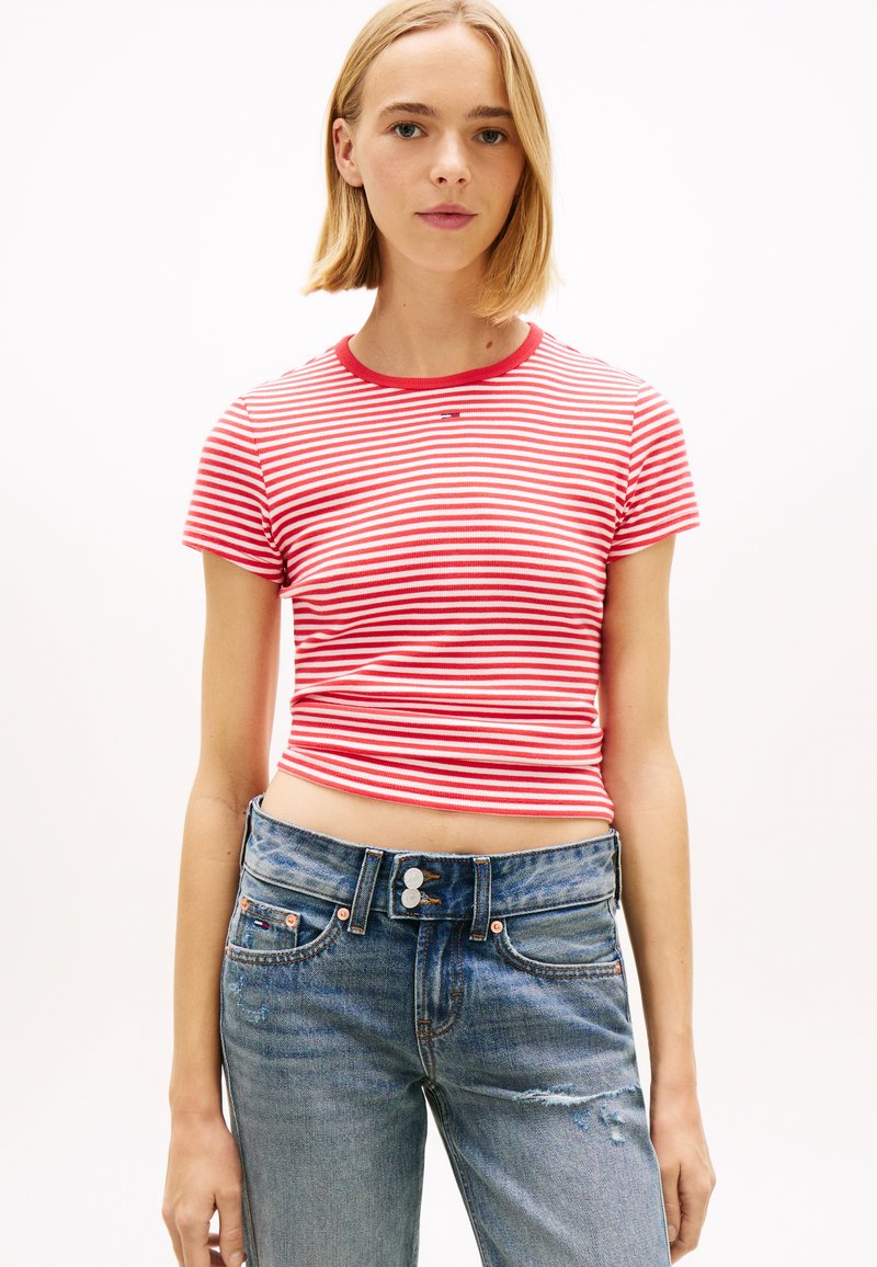 Νέα γυναίκα που φοράει κοντό τοπ με κόκκινες και λευκές ρίγες και μπλε τζιν ψηλόμεσο με φθορές, στέκεται μπροστά σε λευκό φόντο.