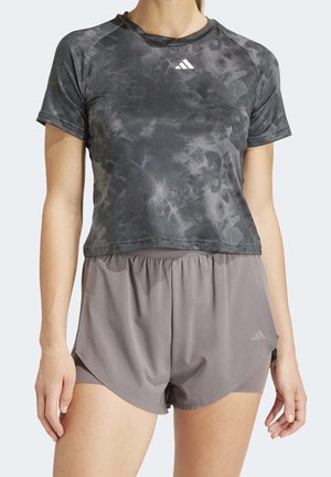 Mujer con una camiseta deportiva corta teñida anudada de color gris oscuro y pantalones cortos de running sueltos en capas de color gris claro, de pie contra un fondo blanco.