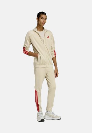 Giovane indossa una tuta beige con strisce rosse sulle maniche e sui pantaloni, abbinata a scarpe da ginnastica bianche, su sfondo chiaro.