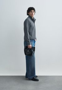 Pruhovaný sveter na zips v sivej a čiernej farbe, modré široké džínsy a čierna kožená kabelka; modelka stojí na sivom podlaží pred bielou stenou.