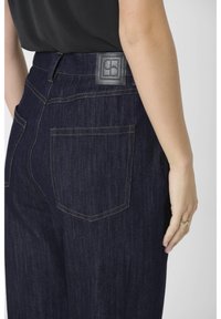 Dark blue denim jeans with a smooth texture, featuring a leather label at the waistband and contrasting yellow stitching on pockets.