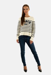 Maglione a righe crema e blu navy con il testo "PARIS", abbinato a jeans scuri e tacchi neri, indossato da un modello su uno sfondo neutro.