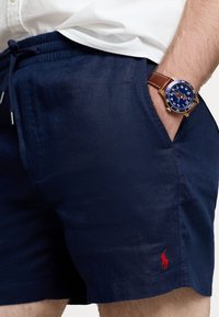 Navy linen swim shorts with an elastic waistband and drawstring. Features a small red embroidered logo and side pockets.