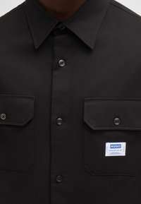 Musta nappitakki, joka on valmistettu puuvillasekoitteesta. Siinä on kaksi rintataskua, kärkikaulus ja merkitty etiketti vasemmassa rintataskussa.