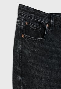Gros plan sur un jean en denim noir montrant la poche avant, la petite poche pour pièces, la boucle de ceinture et les rivets en cuivre sur un fond blanc.