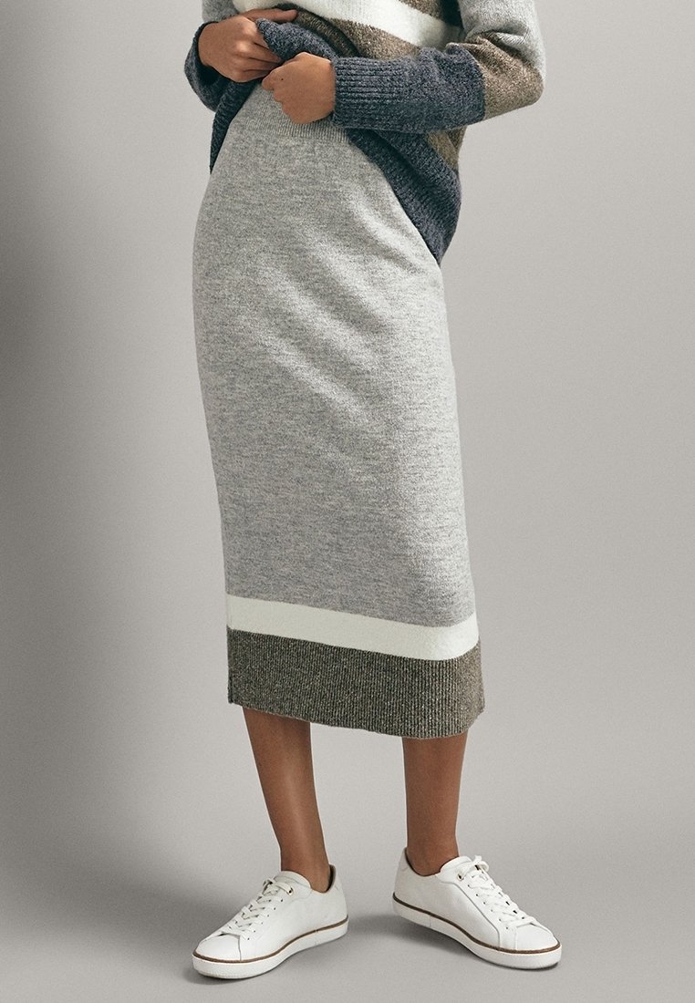Personne portant une jupe midi en maille grise avec des bandes blanches et olive, associée à des baskets blanches et un pull rayé.