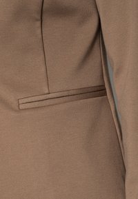 Tissu brun avec une texture lisse, présentant un détail de poche latérale et une coupe ajustée. Le matériau a une légère élasticité et épouse délicatement les formes.