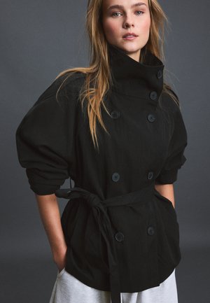 Jeune femme aux longs cheveux portant une veste noire ceinturée à double boutonnage avec des boutons et un pantalon gris clair sur fond gris.