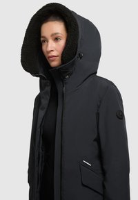 Jachetă puffer neagră cu o glugă mare căptușită cu blană sintetică. Are închidere cu fermoar, buzunare laterale și un accent rotund negru cu logo pe mânecă.