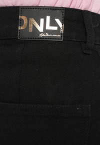 Juodi džinsai su tekstūruota medžiaga, turintys auksiniais akcentais pažymėtą etiketę su užrašu „ONLY“. Juosmens juosta su įprasta džinsų konstrukcija.