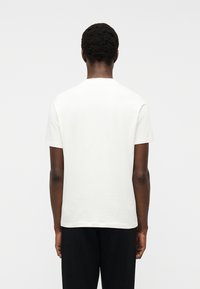 Άνδρας που στέκεται με την πλάτη γυρισμένη, φορώντας απλό λευκό κοντομάνικο μπλουζάκι και μαύρο παντελόνι, μπροστά σε ανοιχτό γκρι φόντο.