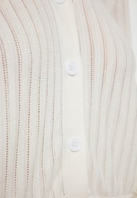 Krémový pletený cardigan se svislými pruhy, s předními knoflíky a kulatými bílými knoflíky na zapínání. Texturovaná, lehká tkanina.