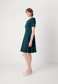 Teal dress with short sleeves, fitted bodice, and flared skirt. Features two decorative buttons on sleeves. Worn with cream slingback heels.