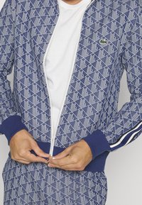 Person som dragit upp en blå mönstrad jacka med vita geometriska former och en liten grön krokodil-logotyp, iförd en vit skjorta under.