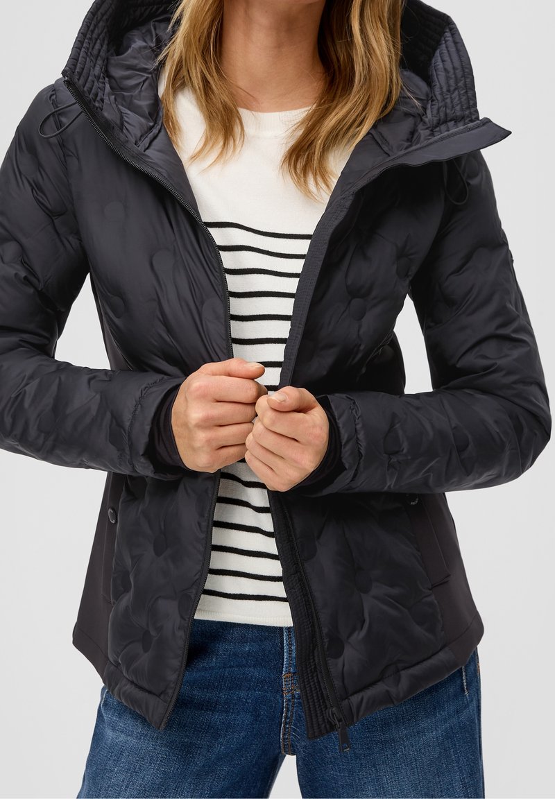 Femme portant une veste matelassée noire par-dessus un pull à rayures blanches et noires, en train de fermer la fermeture éclair de la veste, avec un jean bleu.