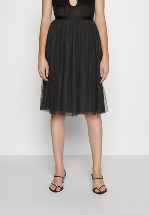 Femme portant une jupe en tulle noire longueur genou avec de petits motifs à pois et des talons noirs ouverts à lanières, debout devant un fond blanc.