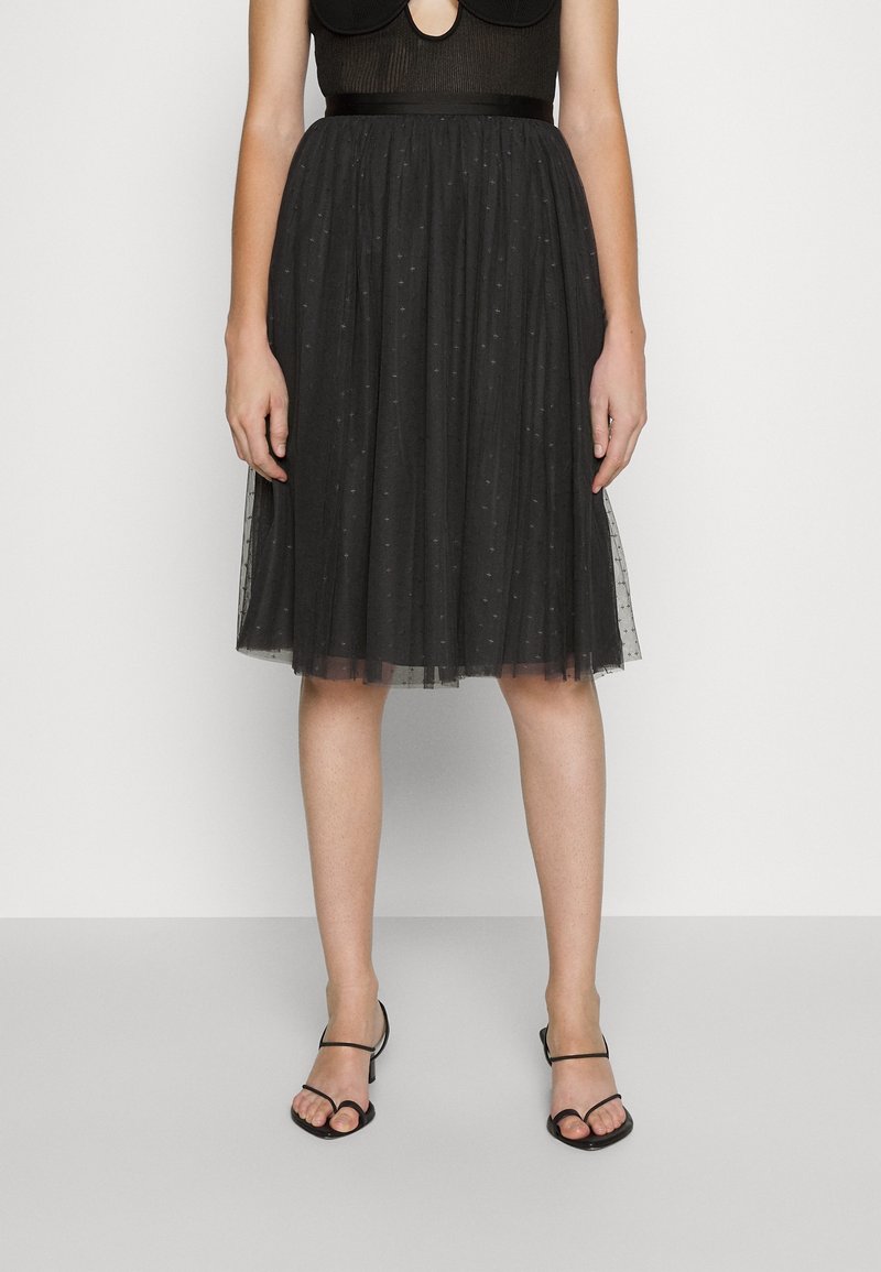 Femme portant une jupe en tulle noire longueur genou avec de petits motifs à pois et des talons noirs ouverts à lanières, debout devant un fond blanc.