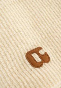 Bonnet en tricot crème avec une texture en côtes, arborant une pièce en cuir marron avec un logo au design stylisé.