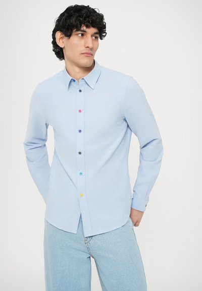 Cămașă cu nasturi bleu deschis, realizată dintr-un material texturat, cu nasturi multicolori pe fanta cu nasturi, asortată cu blugi bleu deschis.