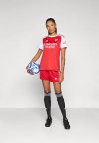 Maillot de football rouge et blanc avec une texture à l'avant, associé à un short rouge. Chaussettes noires avec des rayures blanches et crampons noirs. Tenant un ballon de football multicolore.