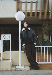 Black leather bomber jacket with white shearling collar, brown wide-leg trousers, patterned accessory, standing near a white lamp post.